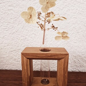 K&ouml;nnte beinhalten: Eine Holzvase mit einem Glas-Reagenzglas zum Halten von Trockenblumen. Die Vase ist aus hellbraunem Holz und hat eine quadratische Form. Das Reagenzglas ist aus klarem Glas und wird in ein Loch in der Mitte der Vase gesteckt. Die Trockenblumen sind wei&szlig; und haben ein zartes, luftiges Aussehen.