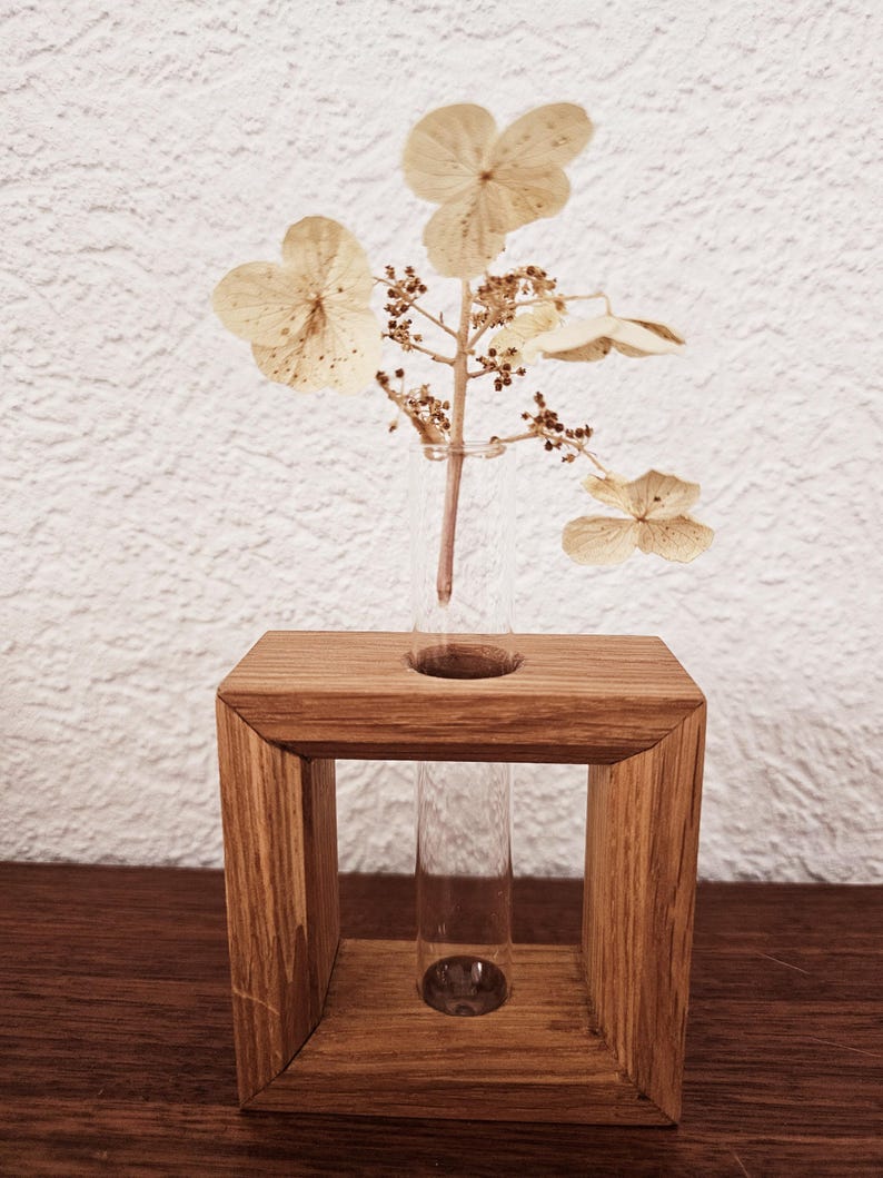 K&ouml;nnte beinhalten: Eine Holzvase mit einem Glas-Reagenzglas zum Halten von Trockenblumen. Die Vase ist aus hellbraunem Holz und hat eine quadratische Form. Das Reagenzglas ist aus klarem Glas und wird in ein Loch in der Mitte der Vase gesteckt. Die Trockenblumen sind wei&szlig; und haben ein zartes, luftiges Aussehen.