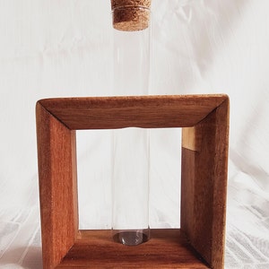 K&ouml;nnte beinhalten: Ein klares Glasr&ouml;hrchen mit einem Korkstopfen sitzt in einem Holzrahmen. Der Rahmen ist aus dunkelbraunem Holz und hat eine quadratische Form.