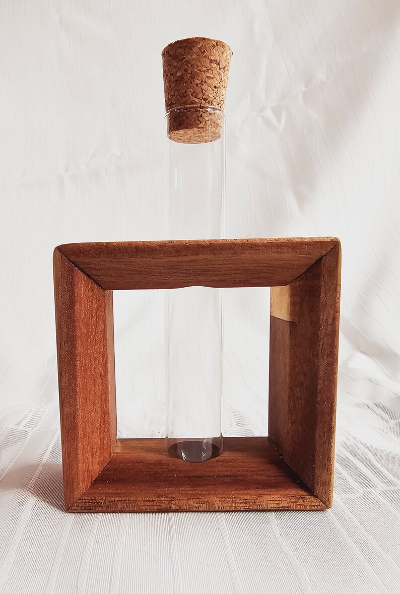 K&ouml;nnte beinhalten: Ein klares Glasr&ouml;hrchen mit einem Korkstopfen sitzt in einem Holzrahmen. Der Rahmen ist aus dunkelbraunem Holz und hat eine quadratische Form.
