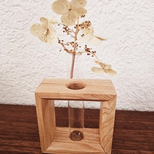 K&ouml;nnte beinhalten: Eine Holzvase mit einem Glas-Reagenzglas-Einsatz, in dem getrocknete wei&szlig;e Blumen stecken. Die Vase ist hellbraun und hat eine quadratische Form.