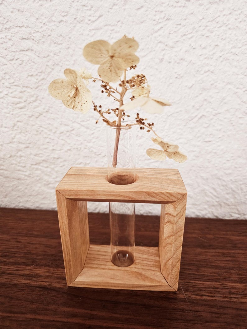 K&ouml;nnte beinhalten: Eine Holzvase mit einem Glas-Reagenzglas-Einsatz, in dem getrocknete wei&szlig;e Blumen stecken. Die Vase ist hellbraun und hat eine quadratische Form.