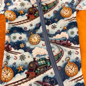 May include: A blue and white baby sleeper with a whimsical train and winter-themed pattern. The design includes trains, pocket watches, snowflakes, and candy canes. The sleeper has a full-length zipper and a light blue trim.