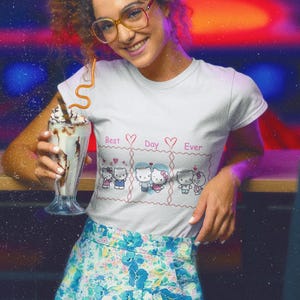 May include: A white t-shirt with a graphic of cartoon cats and the text "Best Day Ever". The person is wearing a floral skirt and holding a milkshake. The background has a blurred, colorful effect.
