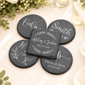 May include: A set of five dark gray slate coasters with rough, natural edges. Four coasters are circular and one is square. Each features white engraved text and botanical designs, including names like Evelyn, Smith, Ashley & Jordan, and Gloria & Brett.