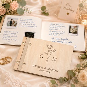 May include: A wooden guest book with a light wood grain finish, featuring the engraved letters G and M, a floral line drawing, and the text Grace & Mason 08.01.2014. The book is open, displaying handwritten notes and small photographs.