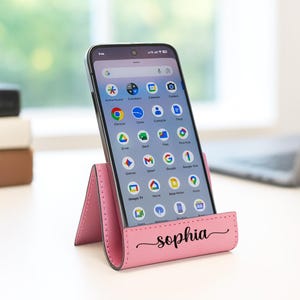 May include: A pink leather phone stand with the name "sophia" in cursive script. A smartphone is displayed in the stand, showing app icons on the screen. The stand is on a white surface.