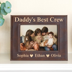 May include: A brown leather picture frame with the words "Daddy's Best Crew" at the top. The frame holds a family photo. Below the photo are the names "Sophia", "Ethan", and "Olivia" with heart symbols.