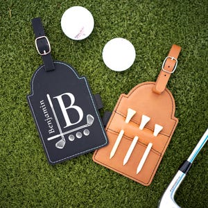 May include: Two personalized golf bag tags, one black and one brown, with white stitching and a leather strap. The black tag has a white golf club graphic and the name "Benjamin" with the letter "B". The brown tag holds golf tees.