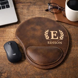 May include: A brown leather mouse pad with a wrist rest, personalized with the name "Edison" and the letter "E" inside a laurel wreath. A black wireless mouse and a laptop are next to the mouse pad.