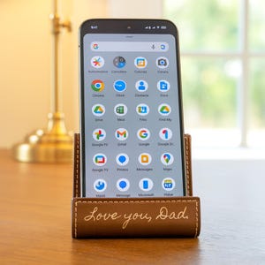Peut inclure: Un smartphone est pos&eacute; dans un support de t&eacute;l&eacute;phone en cuir marron. Le support porte l'inscription "Love you, Dad". L'&eacute;cran du t&eacute;l&eacute;phone affiche diverses ic&ocirc;nes d'applications. Le support est sur une surface en bois.