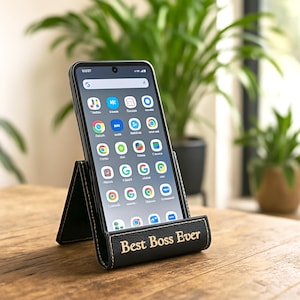 Peut inclure: Un support de t&eacute;l&eacute;phone en cuir noir avec l'inscription dor&eacute;e "Best Boss Ever". Un smartphone est affich&eacute; dans le support, montrant des ic&ocirc;nes d'applications. Le support est sur une surface en bois, avec des plantes vertes en arri&egrave;re-plan.