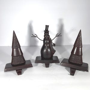 May include: Three dark brown metal Christmas decorations. A snowman with a top hat and scarf is flanked by two Christmas trees. The trees have a textured design. All three decorations have a rectangular base.