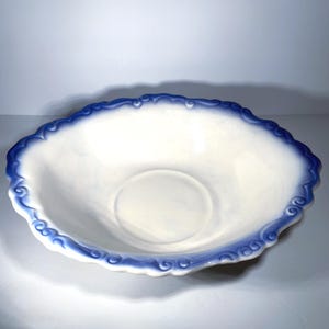 May include: White ceramic serving bowl with a scalloped edge and a blue decorative border. The bowl has a circular indentation in the center. The rim features a repeating scroll design. The bowl is likely for serving food.