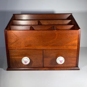 May include: A wooden desk organizer with a rich, dark brown finish. It features multiple compartments for storage and two small drawers with floral-patterned knobs. The organizer is designed to hold various office supplies.