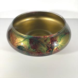 May include: A round, decorative bowl with a gold interior and a colorful exterior. The exterior features a blend of green, red, and gold hues, creating a unique, artistic design. The bowl is made of glass and has a smooth, glossy finish.