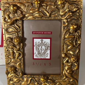 May include: A gold-toned picture frame with a cherub design. The frame has a matted opening with the text "JENNIFER MOORE" and "3 1/2 X 5" printed in red.