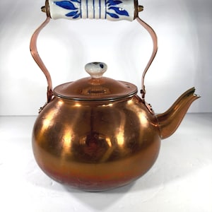 May include: A copper tea kettle with a round body and a curved spout. The handle is white ceramic with blue floral designs. The lid has a small ceramic knob. The kettle is a vintage style.