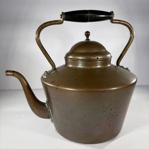 May include: Antique copper kettle with a black wooden handle. The kettle has a spout and a lid with a small knob. The handle is attached to the body with brass brackets. The kettle has a vintage look and feel.