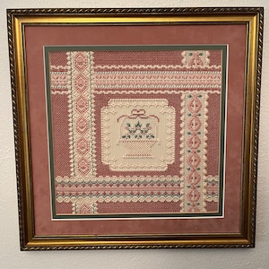 May include: A framed cross-stitch embroidery with a pink and white floral design on a red background. The embroidery is surrounded by a gold frame.