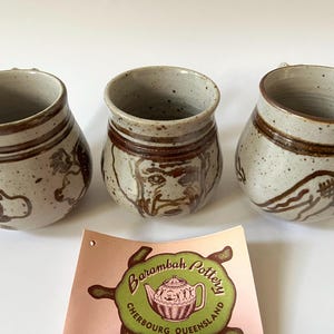 May include: Three speckled ceramic mugs with brown painted designs. Each mug has a unique illustration, including a bird, a face, and an abstract design. The mugs have a handle and a brown band around the top.