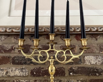 May include: A gold metal candelabra with five arms, each holding a black candle. The candelabra has a decorative, ornate design and sits on a brick surface.