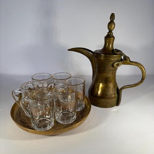 May include: A set of six clear glass cups with gold trim, a brass tray with intricate designs, and a brass teapot with a hinged lid and a spout. The teapot has a decorative finial on top.