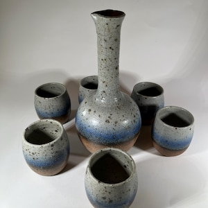 May include: A set of six ceramic drinking vessels, including a tall decanter with a wide base and five smaller cups. The pieces are all glazed in a speckled white with a blue and brown band around the middle.