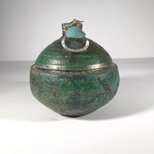 May include: A handcrafted, green ceramic jar with a lid. The jar has a textured surface and a metallic rim. The lid features a small, turquoise-colored accent and a silver ring. The jar is a unique decorative item.