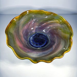 May include: A decorative glass bowl with a swirling design. The bowl features a deep blue center, transitioning to shades of purple, green, and pink. The rim is edged with a gold color, and the bowl has a wavy, organic shape.