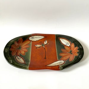 May include: An oval ceramic serving dish with a green and orange glaze. The dish is decorated with a dragonfly and stylized flowers.