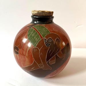 May include: A round, brown decorative bottle with a monkey design. The bottle has a dark brown base and a light brown wooden stopper. The monkey is etched in a lighter brown color, with a green leaf in the background.