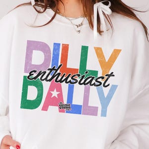 May include: White sweatshirt with the text "DILLY enthusiast DALLY" in colorful block letters. The sweatshirt has a crew neck and long sleeves. The Cosmic Studio shop logo is visible.