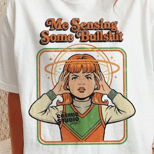 Peut inclure: T-shirt blanc avec l'inscription "Me Sensing Some Bullshit" au-dessus d'une illustration rétro d'une femme les mains sur la tête, dans un cadre vert et orange. Le logo Cosmic Studio Shop est également visible.