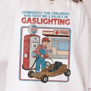 以下が含まれることがあります： 「CURRENTLY THE CHEAPEST GAS THAT WE CAN GET IS GASLIGHTING」と書かれたグラフィックが特徴の白いスウェットシャツ。子供と木製のゴーカートが登場するレトロなガソリンスタンドのシーンが描かれています。Cosmic Studio shopのロゴが左上にあります。