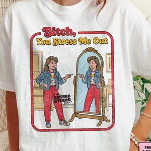 以下が含まれることがあります： レトロなグラフィックの白いTシャツ。鏡に映った自分の姿を指さす女性が描かれており、「Bitch, You Stress Me Out」というテキストが入っています。デザインは赤い縁で囲まれ、Cosmic Studio shopのロゴが入っています。