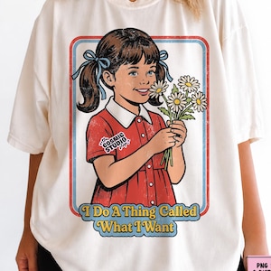 Op de afbeelding: Crèmekleurig T-shirt met een retro-illustratie van een meisje in een rode jurk met madeliefjes. De afbeelding is omlijst met een rode en blauwe rand en bevat de tekst "I Do A Thing Called What I Want".