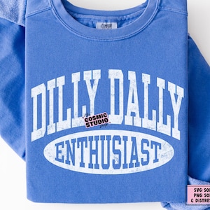 K&ouml;nnte beinhalten: Ein blaues Sweatshirt mit Rundhalsausschnitt und wei&szlig;em, abgenutztem Text, der DILLY DALLY ENTHUSIAST in einem College-Stil zeigt. Das Design enth&auml;lt eine ovale Form um das Wort ENTHUSIAST und ein kleines Logo f&uuml;r The Cosmic Studio Shop.