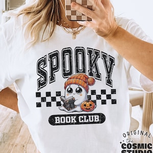 May include: A white t-shirt featuring a black and white graphic of a ghost wearing a hat and holding a book. The text "Spooky Book Club" is printed below the ghost. The text "the COSMIC STUDIO shop" is printed in the bottom left corner of the image. The text "ORIGINAL the COSMIC STUDIO shop DESIGNER" is printed in the bottom right corner of the image. The text "PNG SOLID & DISTRESSED" is printed at the bottom centre of the image.