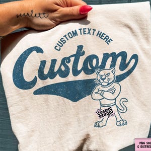 Może przedstawiać: Kremowy t-shirt z niebieską grafiką o postarzanym wyglądzie. Wzór zawiera napis "CUSTOM TEXT HERE", słowo "Custom" dużą czcionką skryptową oraz ilustrację pantery ze skrzyżowanymi ramionami na klatce piersiowej.