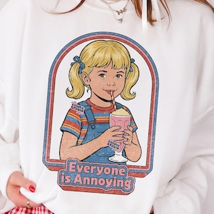 Könnte beinhalten: Weißes Sweatshirt mit einer Retro-Illustration eines Mädchens, das einen Milchshake trinkt, mit dem Text "Everyone is Annoying". Das Design ist im Vintage-Stil gerahmt. Das Logo des Cosmic Studio Shops befindet sich oben links.