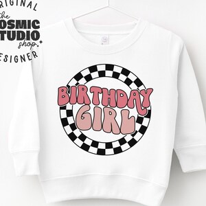 May include: White sweatshirt with a black and white checkered circle design. The text "BIRTHDAY GIRL" is in pink and white letters inside the circle.