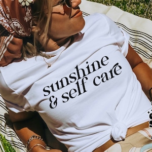 May include: A white t-shirt with black text that reads "sunshine & self care". The shirt is worn by a person lying on a striped blanket. The image includes a logo for "the cosmic studio shop" in the bottom left corner and a logo for "the cosmic studio shop designer" in the bottom right corner.