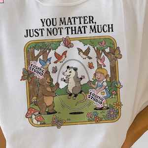 Puede incluir: Camiseta blanca con la frase "YOU MATTER, JUST NOT THAT MUCH" y una ilustración de dibujos animados. El diseño presenta un oso, una zarigüeya y una niña jugando a la comba en un bosque con mariposas y pájaros. La camiseta también tiene el texto "the COSMIC STUDIO shop."