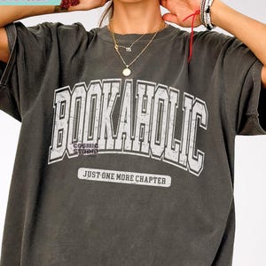 Puede incluir: Una camiseta gris con un gráfico blanco que dice "BOOKAHOLIC" en una fuente de estilo universitario vintage. El gráfico también incluye el texto "JUST ONE MORE CHAPTER".