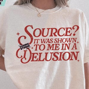 Op de afbeelding: Crèmekleurig T-shirt met de rode tekst "SOURCE? IT WAS SHOWN TO ME IN A DELUSION". Het shirt heeft ook de tekst "COSMIC STUDIO shop." Het ontwerp is een digitale download.