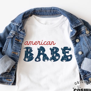 May include: White t-shirt with the text "american BABE" in blue and red letters with stars. The text is distressed and has a vintage look. The t-shirt is on top of a blue denim jacket. The text "the COSMIC STUDIO shop. ORIGINAL DESIGNER" is in a circle at the bottom of the image.
