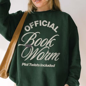 Può includere: Una felpa verde scuro con la scritta bianca "OFFICIAL Book Worm" e "Plot Twists Included". Il design è solido e usurato, parte di un download digitale, disponibile in tre colori diversi.