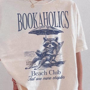 May include: A white t-shirt featuring a raccoon graphic and the text "Bookaholics Beach Club Just one more chapter".