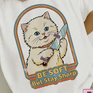 May include: White t-shirt featuring a cartoon cat holding a knife within a retro-style frame. The design includes the text "BE SOFT But Stay Sharp" in a playful font. The shirt has a distressed look.